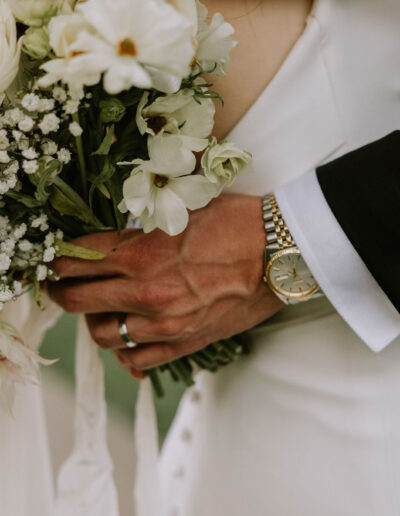 Close-up of a couple holding a bouquet of white and peach flowers. The person on the right wears a black suit and a watch.