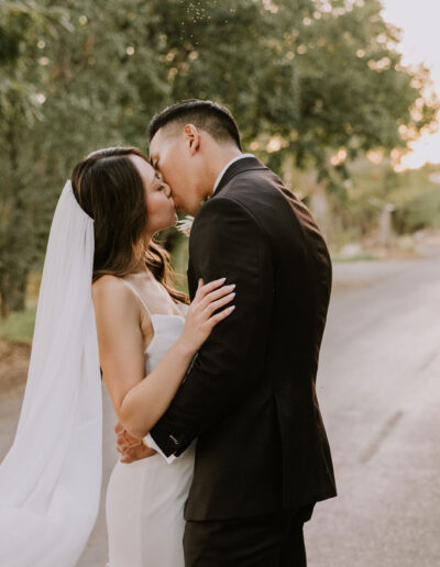 A couple in wedding attire shares a kiss on a tree-lined road at sunset.