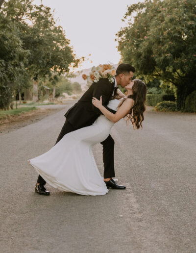 A couple in formal attire embraces in a dip on a tree-lined road, with the woman holding a bouquet of flowers.