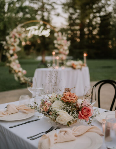 Elegant outdoor wedding table setting with floral centerpiece, candles, and a decorative arch in the background.