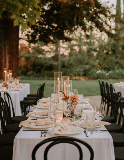 Elegant outdoor wedding setup with round tables covered in white tablecloths, black chairs, floral centerpieces, and tall candle arrangements in a lush garden setting.