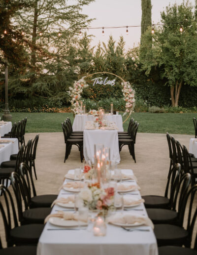 Outdoor wedding reception setup with long tables covered in white tablecloths, black chairs, floral centerpieces, and a decorative arch with the text "The Lees" in the background.