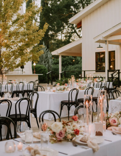 Outdoor wedding reception with elegantly set tables, white tablecloths, floral centerpieces, and black chairs. String lights hang above, creating a warm ambiance near a white building and trees.