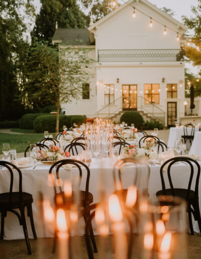 Outdoor wedding reception setup with long tables, white tablecloths, floral centerpieces, candles, and black chairs. A white house and string lights are visible in the background.