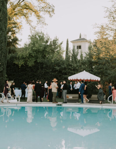 People gathered around a pool with trees and a building in the background on a clear day.