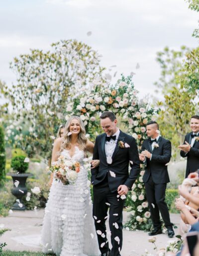 A bride and groom walk down the aisle outdoors, surrounded by guests throwing flower petals. An arch decorated with flowers is in the background.