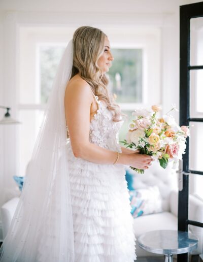 Bride in a white gown holding a bouquet stands by a window, with long veil and tiered dress facing left.