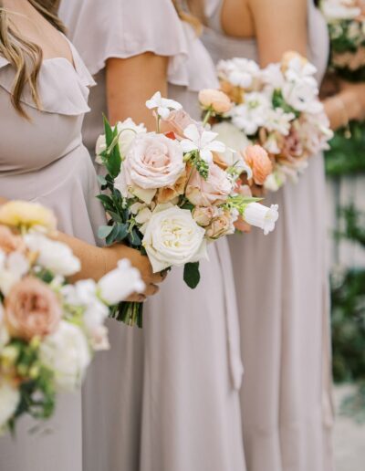 Bridesmaids in light dresses holding bouquets of roses and other flowers.