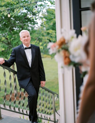 Man in a black tuxedo stands on stairs, holding a railing, with a blurred bouquet in the foreground. Outdoor greenery is visible in the background.