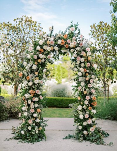 An archway decorated with lush green foliage and pastel roses, set outdoors with trees and a garden visible in the background.