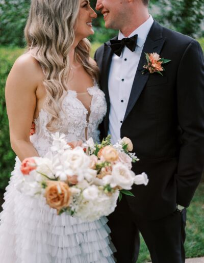 A bride and groom smiling at each other, holding a bouquet of white and peach flowers. The bride is in a white dress, and the groom is in a black tuxedo with a boutonniere.