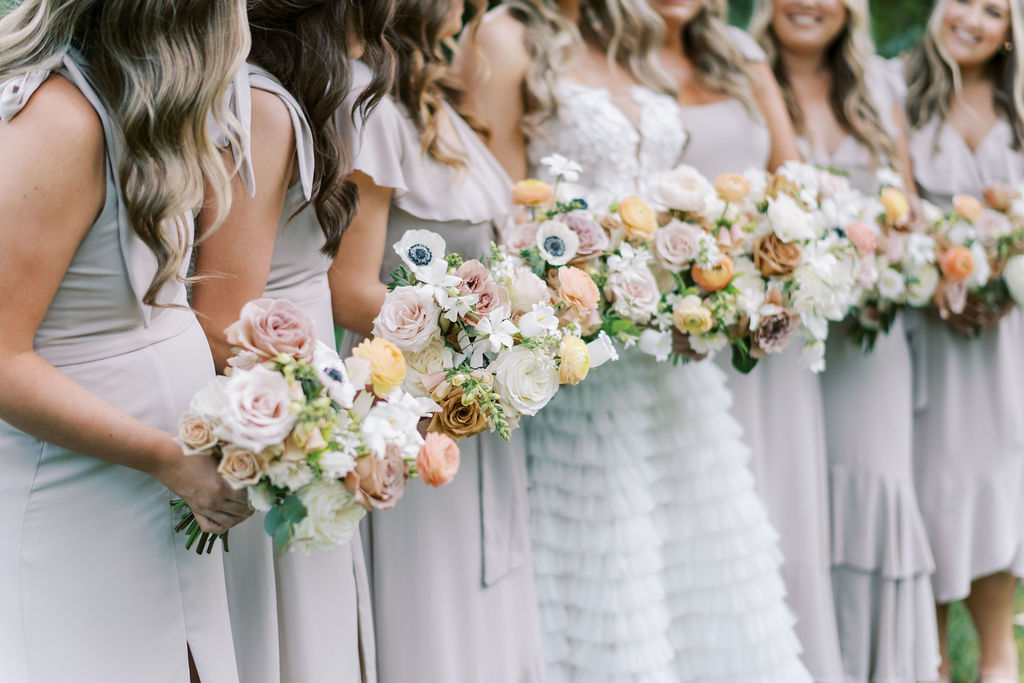 At Park Winters, bridesmaids in light beige dresses holding pastel flower bouquets are perfectly poised beside a radiant bride in a white gown.