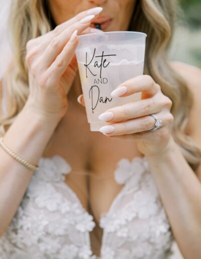 Woman in a white floral dress drinking from a clear cup with "Kate and Dan" written on it.