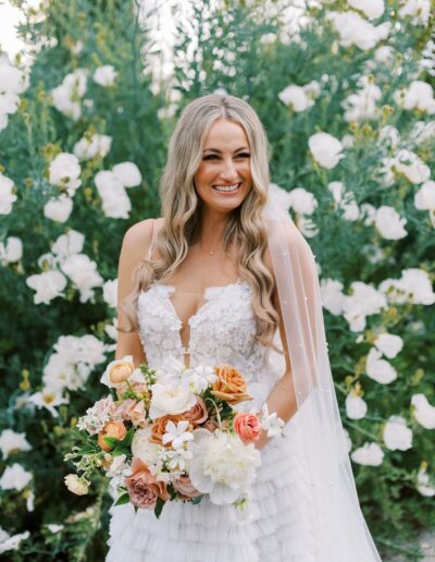 A bride with long wavy hair smiles, holding a bouquet of white, orange, and pink flowers. She stands in front of a backdrop of white flowering plants, wearing a detailed white wedding dress.