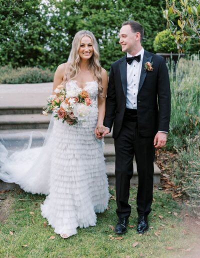 A bride in a white dress and a groom in a black tuxedo hold hands and smile in a garden setting. She holds a bouquet of flowers.