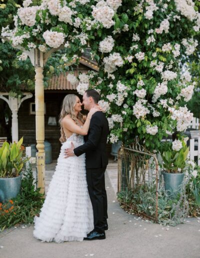 A bride and groom embrace under an arch covered with white flowers in an outdoor setting.