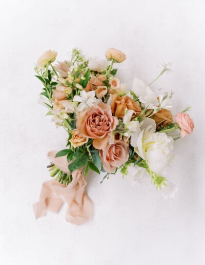 A bouquet of flowers featuring peach, pink, and white blooms with greenery and a light pink ribbon on a white background.