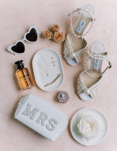 Flat lay of bridal accessories: pearl-embellished shoes, sunglasses, perfume, jewelry, and a white bag with "MRS" on it, surrounded by roses and a white flower.