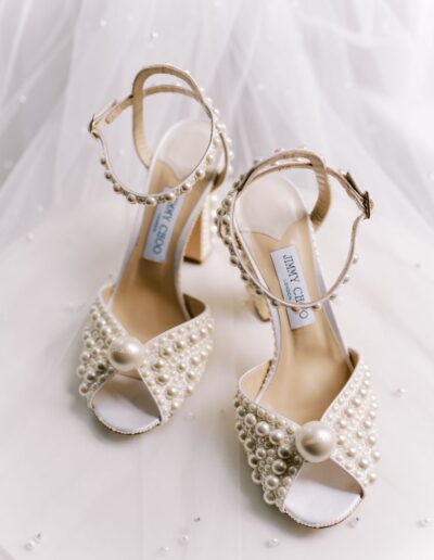 Beige high-heeled sandals adorned with pearl embellishments and ankle straps, set on a white fabric background.