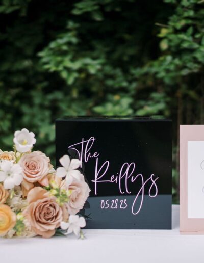 A black box with "The Kellys 05.08.23" next to a card and floral arrangement, set against green foliage.