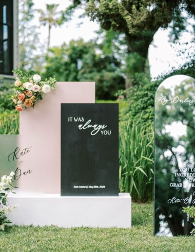 Floral signs outdoors read "IT WAS always YOU" and "Hey Cutie, you look incredible. Now go grab a drink." Surrounded by lush greenery and floral arrangements.