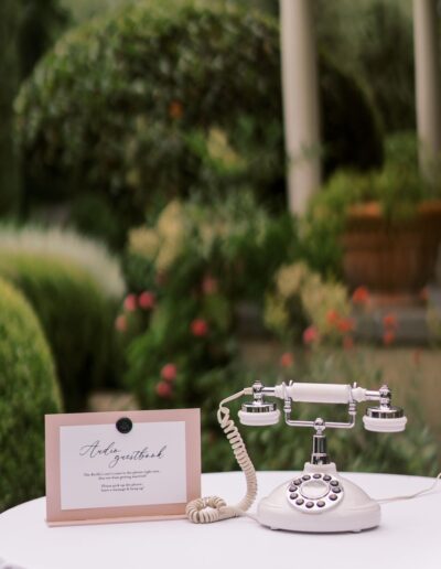 A vintage rotary telephone sits on a table outdoors next to a guestbook sign, with plants and a columned structure in the background.