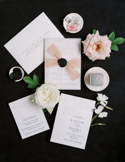 A wedding invitation suite with engraved details, floral embellishments, and wax seals is arranged on a dark surface alongside flowers and a vintage-style stamp.