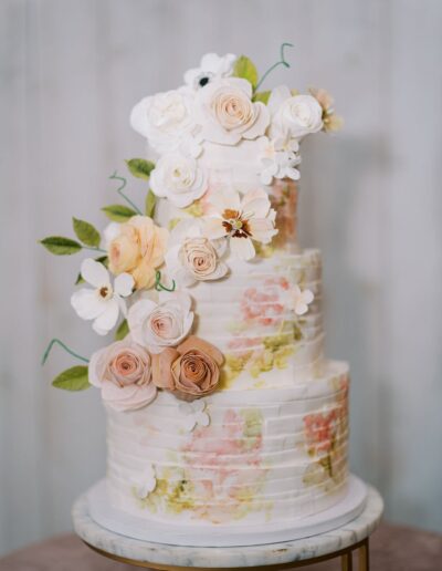 A three-tiered wedding cake with pastel floral decorations and leaves, placed on a round marble stand.