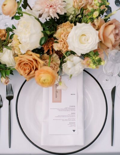 Elegant table setting with floral arrangements in peach and white tones, featuring a menu placed on a white plate with black cutlery on either side.