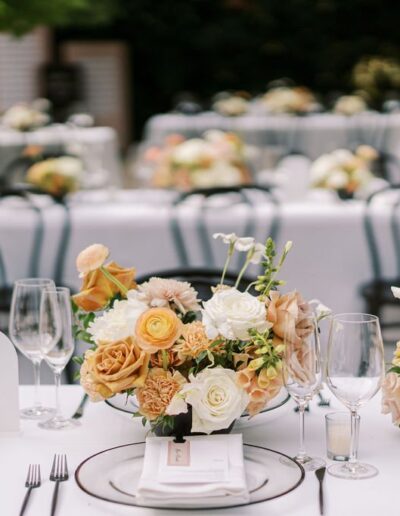 Elegant outdoor dining setup with white tablecloths, floral centerpieces featuring roses, and neatly arranged cutlery and glassware. Black chairs surround the tables.