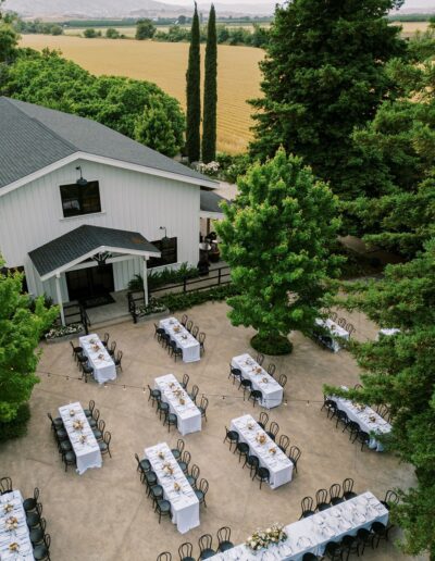 An outdoor wedding setup with rows of tables and chairs arranged on a patio near a white building, surrounded by trees and fields.