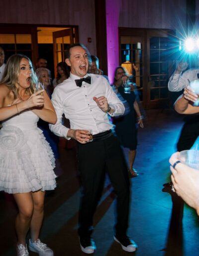 People in formal attire dance and hold drinks at a lively indoor party, with colorful lighting and a bright flash in the background.