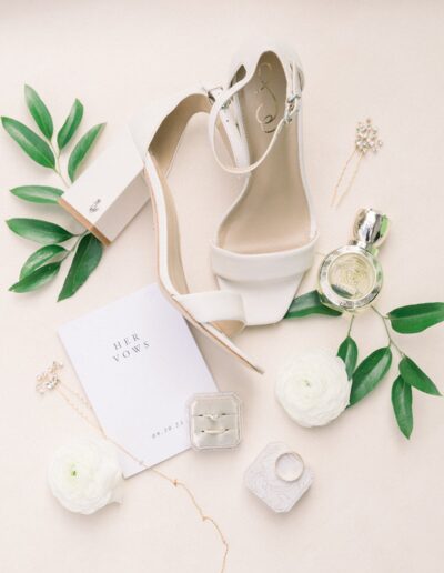 Flat lay of wedding accessories: white high-heeled shoes, perfume, ring, floral decoration, vow booklet with "HER VOWS 09.30.23," and a small box on a light background.