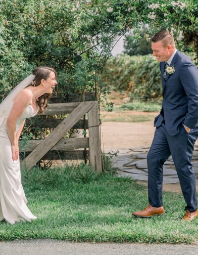 A bride in a white dress and veil laughs while leaning forward. A groom in a blue suit and brown shoes stands facing her, smiling outdoors near greenery and a wooden gate.