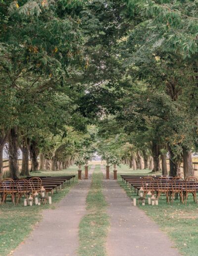 Outdoor wedding ceremony setup with wooden chairs and benches arranged in rows beneath a canopy of trees. Candles in glass holders line the aisle on a path.