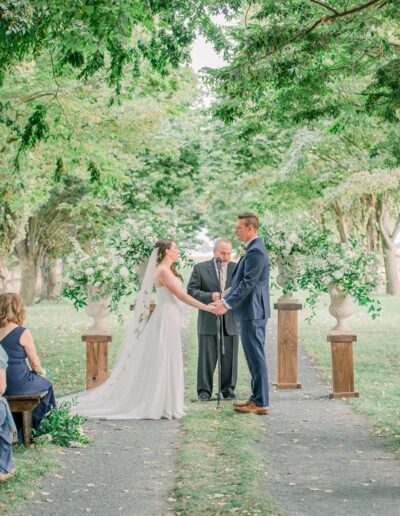 A couple stands at an outdoor wedding ceremony, holding hands in front of an officiant. They are surrounded by greenery and floral arrangements.