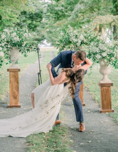 A couple kisses on a tree-lined path, with floral arrangements on pedestals in the background. The bride's dress has a long train.