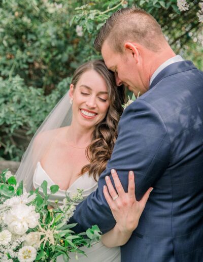 A bride and groom embrace outdoors with greenery and white flowers. The bride smiles with her eyes closed, and the groom wears a dark suit.