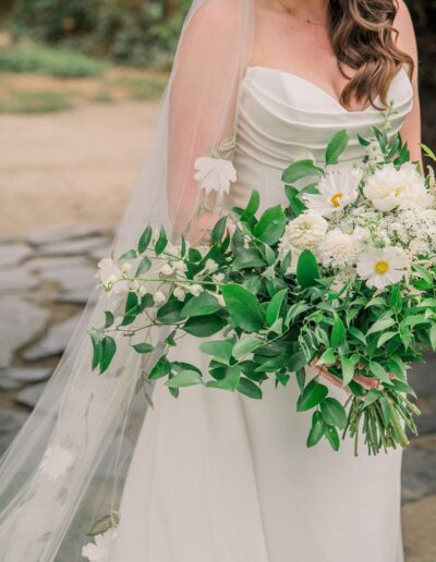 Bride holding a bouquet of white flowers and green leaves, wearing a white strapless gown with a long veil adorned with flowers. Background features stone path and greenery.