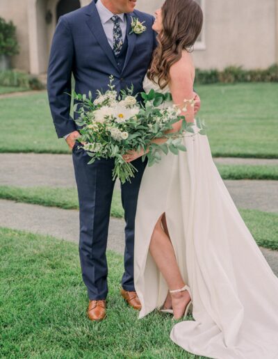 Bride and groom stand on grass, holding a bouquet of flowers. Bride wears a white dress with a slit, groom in a navy suit. House in the background.