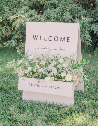 Welcome sign on grass with white flowers in front, reads "We’re glad you’re here, Kristin and Travis." Surrounded by greenery.