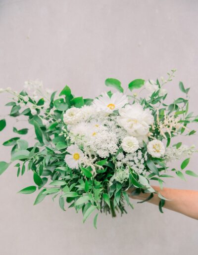 A hand holding a bouquet of white flowers and greenery against a plain background.