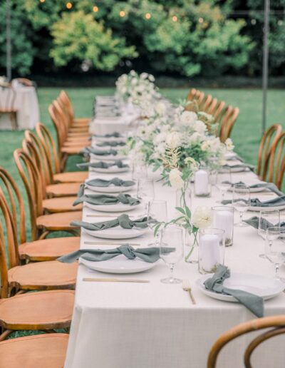 An elegantly set outdoor dining table with wooden chairs, white tablecloth, green napkins, floral centerpieces, and glassware. Lush greenery and string lights in the background.