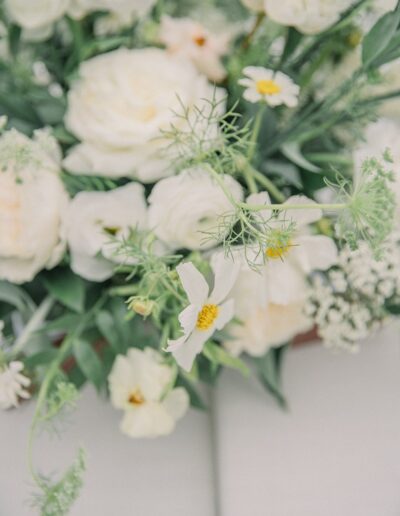 A close-up of a floral arrangement featuring white roses and delicate white daisies with yellow centers, surrounded by green foliage on a light background.