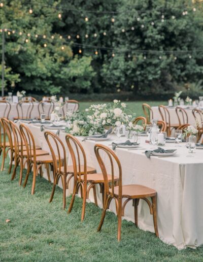 Outdoor wedding reception setup with long tables covered in white tablecloths, wooden chairs, floral centerpieces, and string lights overhead.