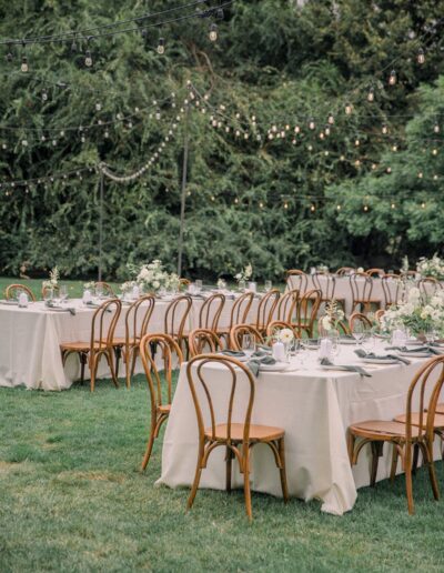 Outdoor event with long tables and wooden chairs set on grass. Tables are covered with white cloths, decorated with flowers. String lights are hung above, surrounded by trees.