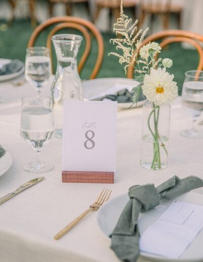 Dining table set for an event with a table number 8 card, glassware, white plates, green napkins, and a small floral arrangement in a glass vase.