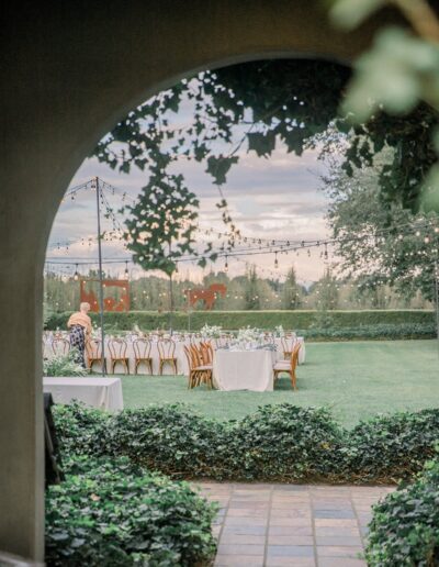 Outdoor event space with decorated tables and chairs under string lights, viewed through an archway with greenery.