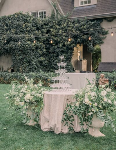 Champagne glass tower on a draped table surrounded by white flowers and greenery, placed outdoors in front of an ivy-covered building with string lights above.