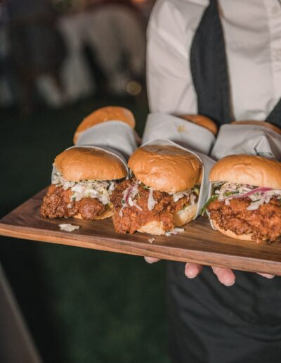 A person holds a wooden tray with multiple fried chicken sandwiches, each topped with coleslaw and wrapped partially in white paper.